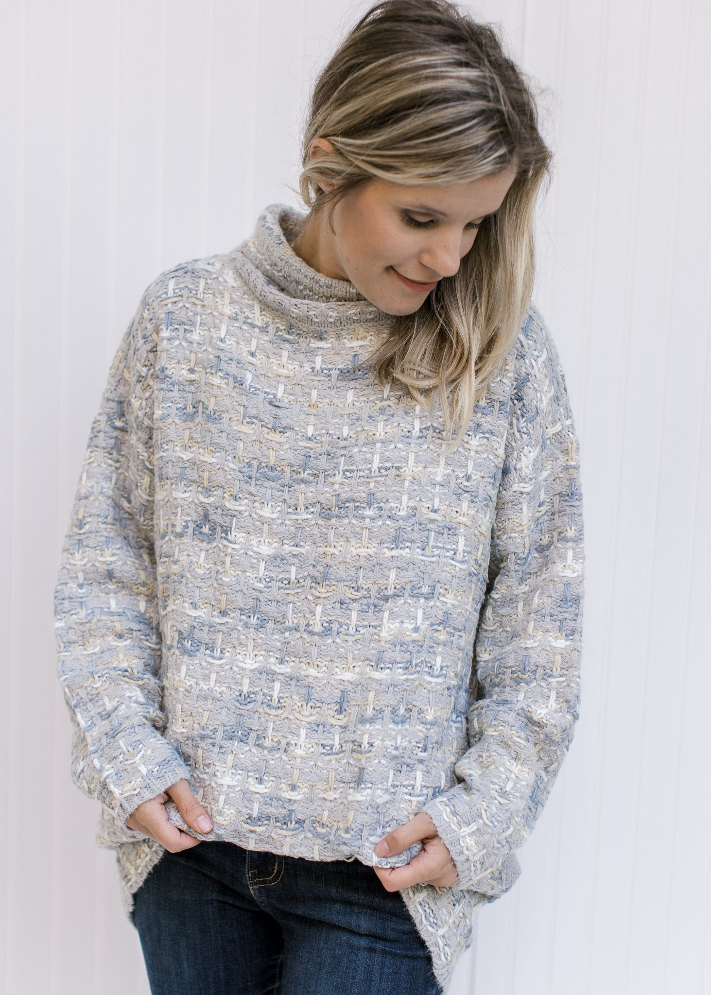 Model wearing a long sleeve gray, cream and mustard tweed sweater with a turtleneck and pockets. 