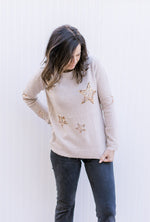 Model wearing black jeans and a taupe sweater with gold sequin stars and long sleeves.