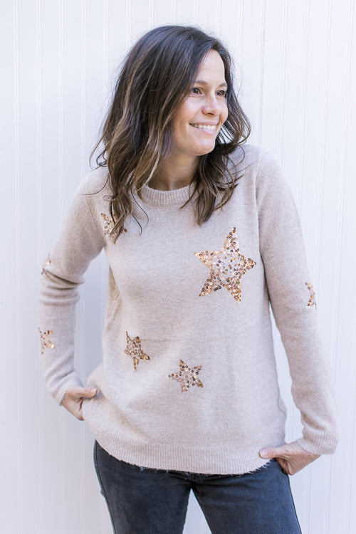 Model wearing a taupe sweater with gold sequin stars, long sleeves and a round neck.