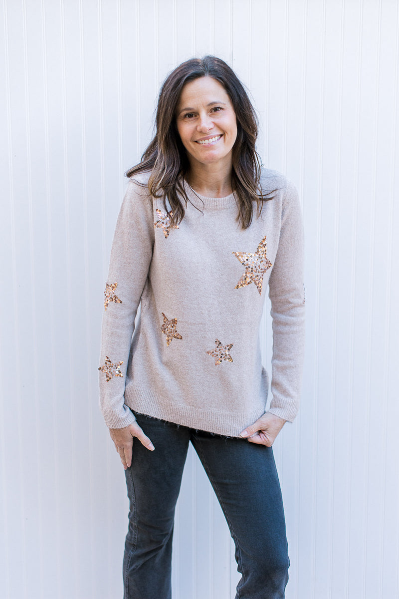 Model wearing a long sleeve taupe sweater with gold sequin stars and a round neck.