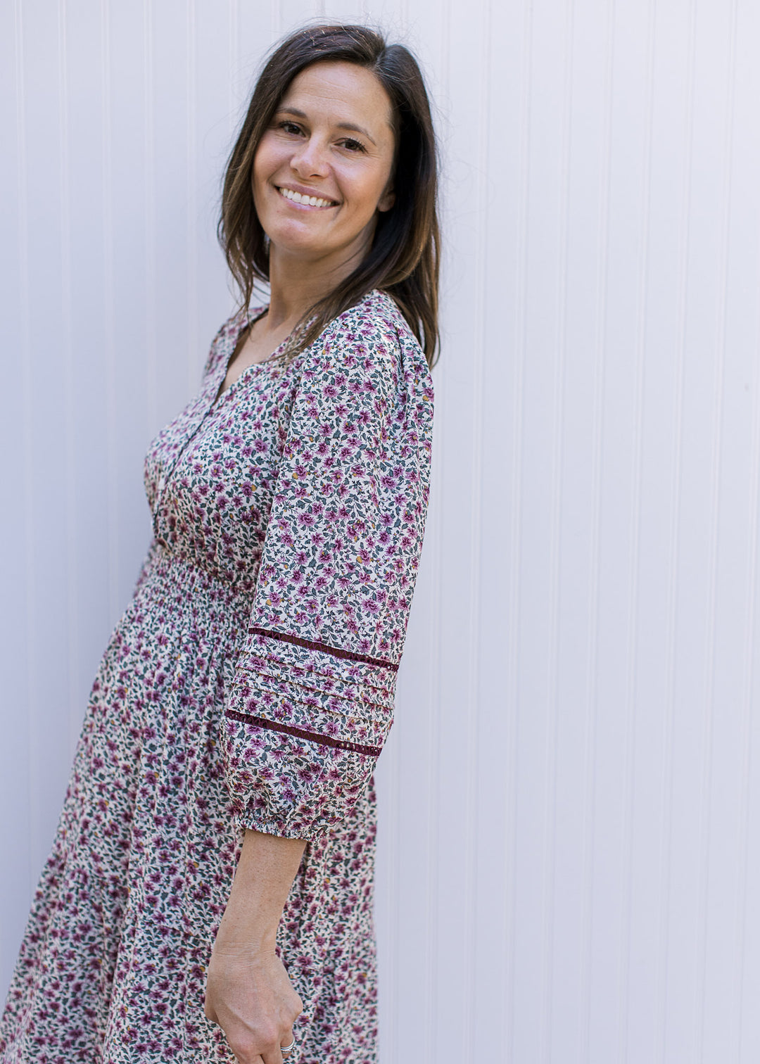 Model wearing a cream v-neck dress with plum ditsy flowers, 3/4 sleeve and a smocked waist. 