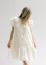 Model wearing a cream above the knee dress with pockets, ruffle cap sleeve and button down front. 