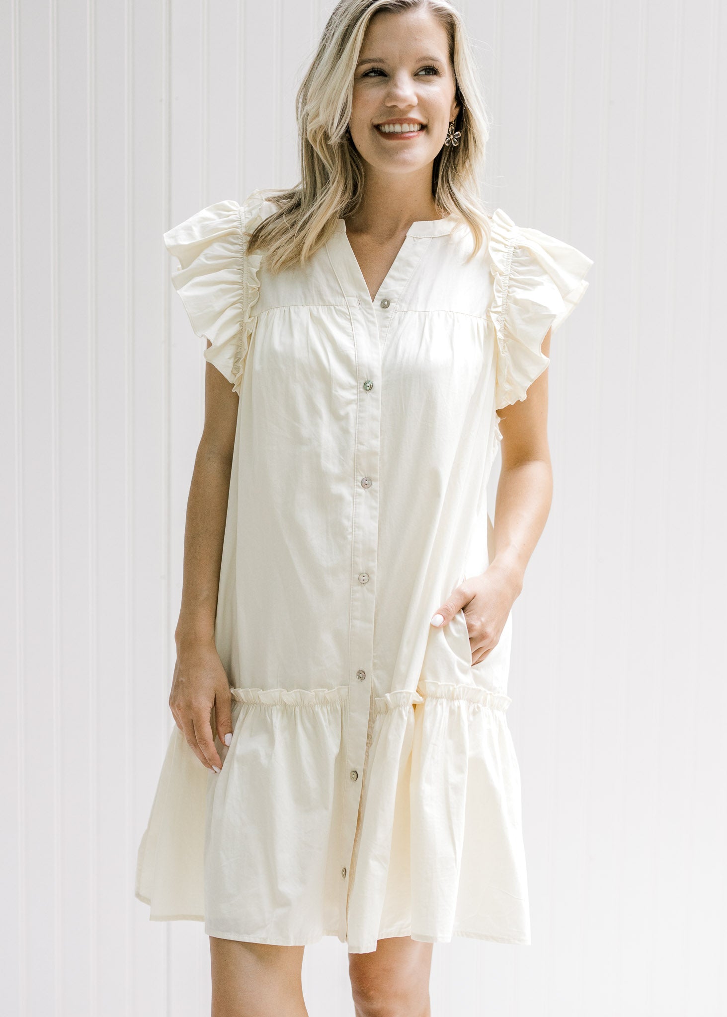 Model wearing a cream dress with pockets, ruffle cap sleeve and button down front. 