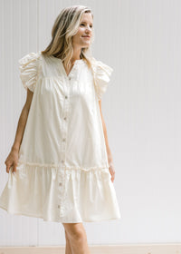 Model wearing a cream dress with pockets, ruffle cap sleeve and button down front and ruffle at base