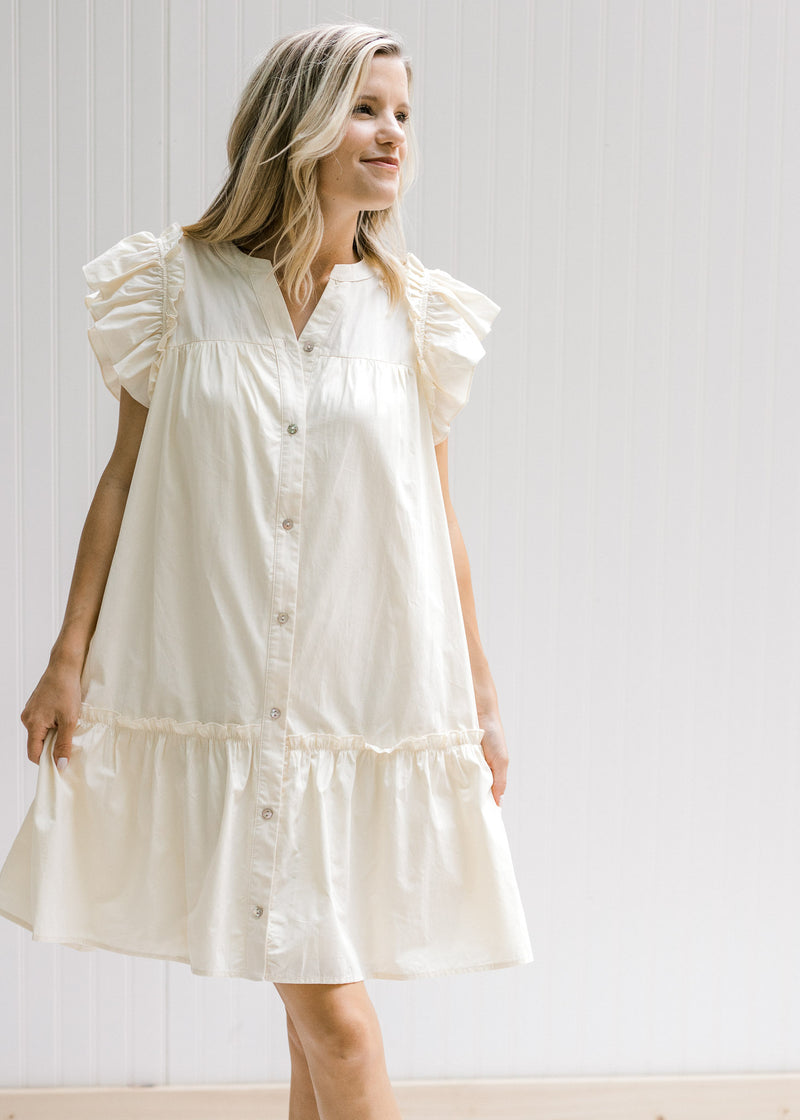 Model wearing a cream dress with pockets, ruffle cap sleeve and button down front and ruffle at base