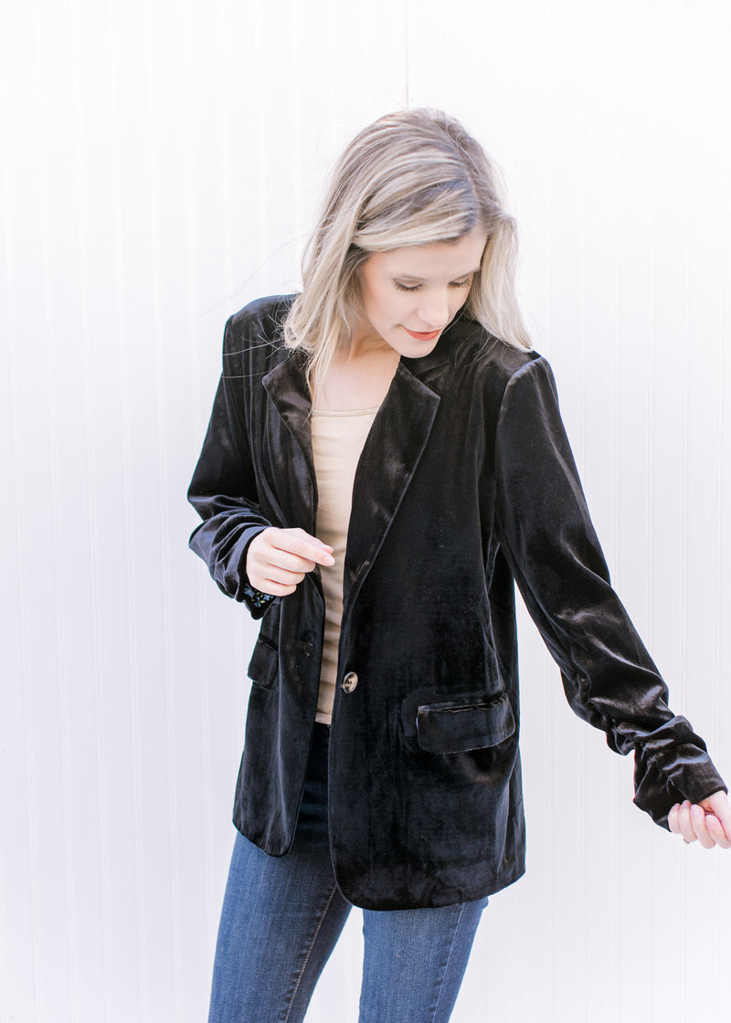 Model wearing a black velvet blazer with faux flap pockets and long sleeves with a ruched detail. 