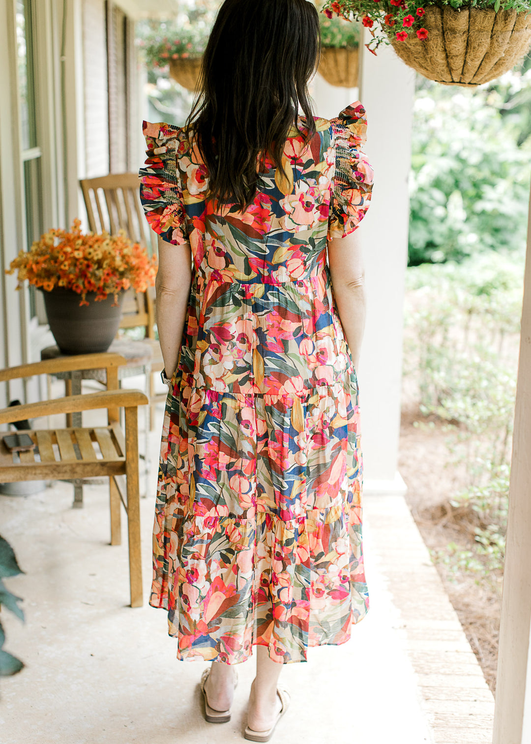 Back view of Model wearing a bright floral midi with cap sleeves, round neck and a tiered skirt.