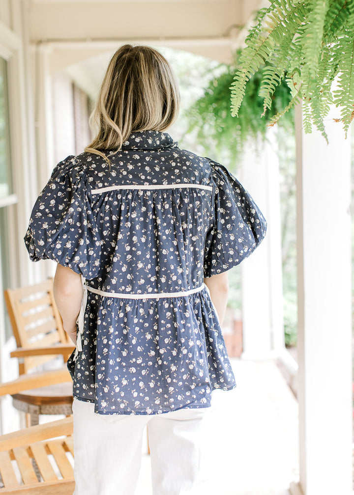 Back view of Model wearing a navy top with white flowers, short puff sleeves and a side tie detail.