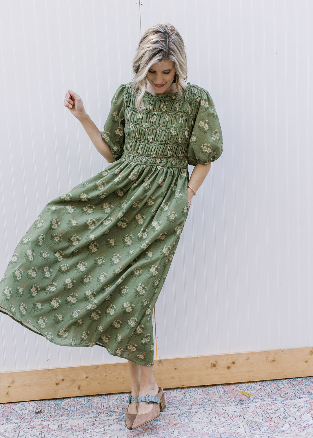 Model wearing heels and an olive midi with cream floral, smocked bodice and elbow-length sleeves. 