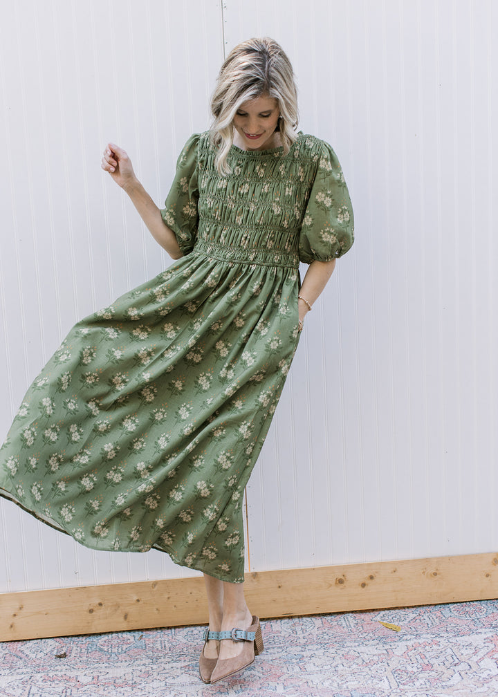 Model wearing heels and an olive midi with cream floral, smocked bodice and elbow-length sleeves. 