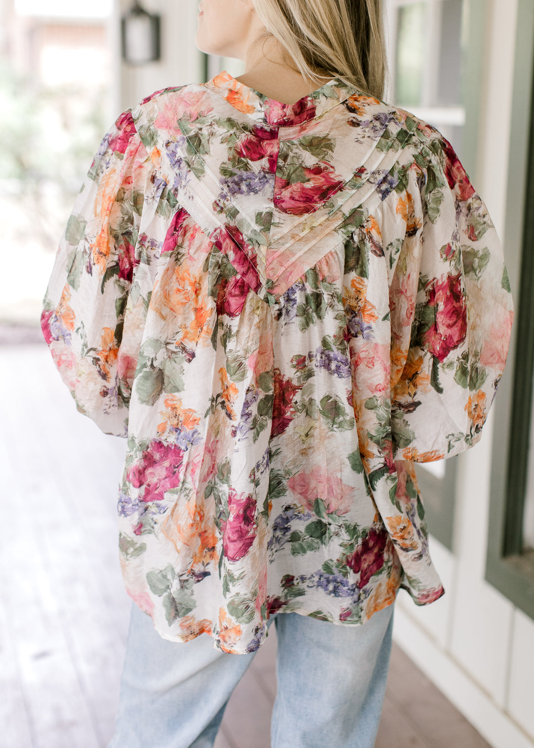 Back view of Model wearing a floral long sleeve top with covered buttons and wide cuffs. 
