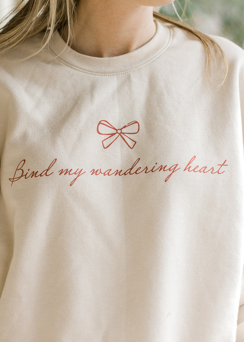 Close up of red script “bind my wandering heart” and bow on a cream sweatshirt with round neck. 