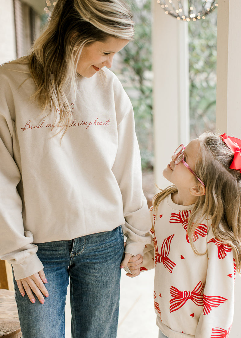 Model, with child, wearing a cream long sleeve sweatshirt with red “bind my wandering heart” script.