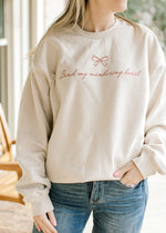 Model wearing a cream sweatshirt with red “bind my wandering heart” script and long sleeves. 