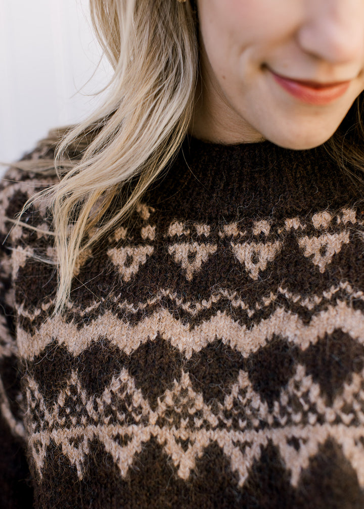 Close up of tan geometric pattern on a brown sweater with a round neck and long sleeves. 