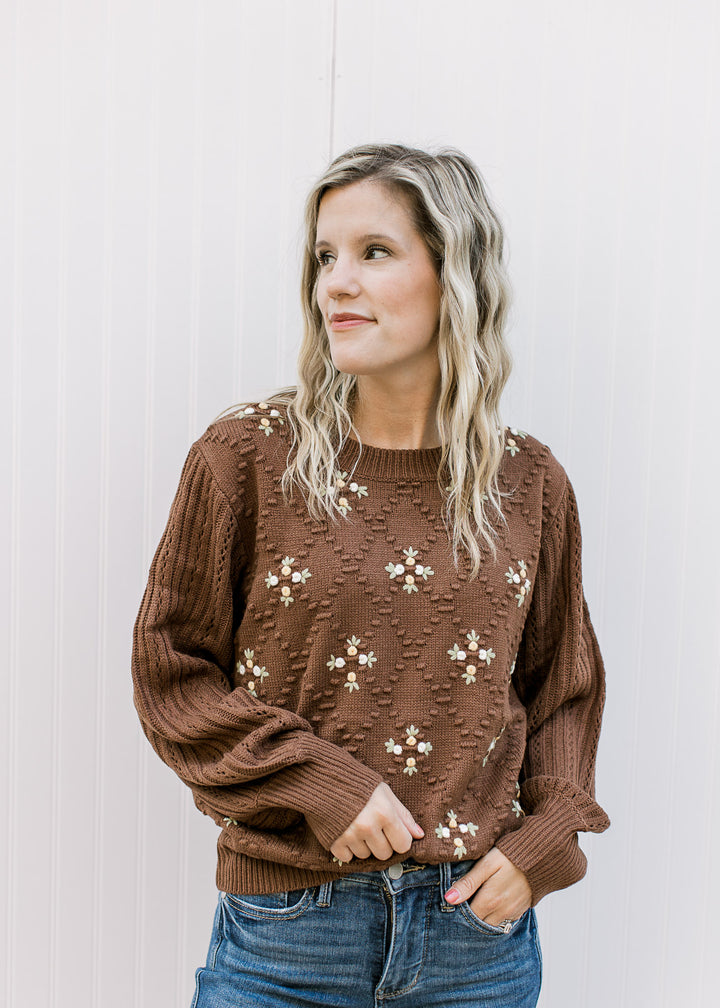 Model wearing a long sleeve mocha brown sweater with a diamond knit pattern and floral accents. 