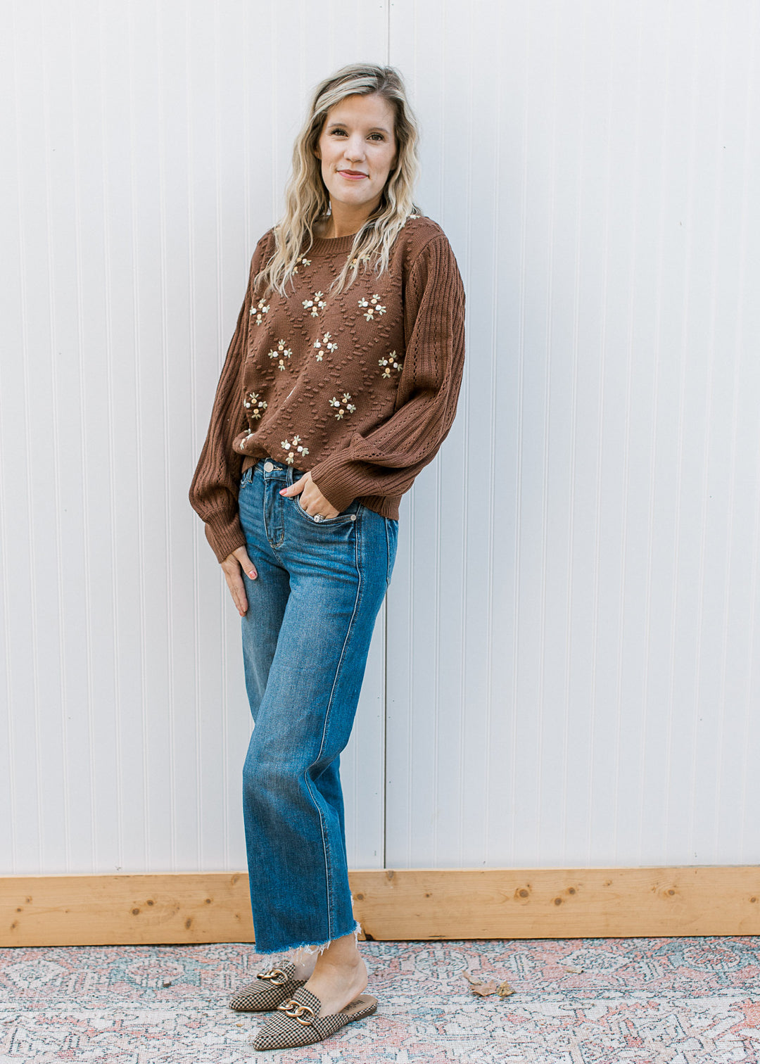 Model wearing jeans and a long sleeve mocha sweater with a diamond knit pattern and floral accents. 