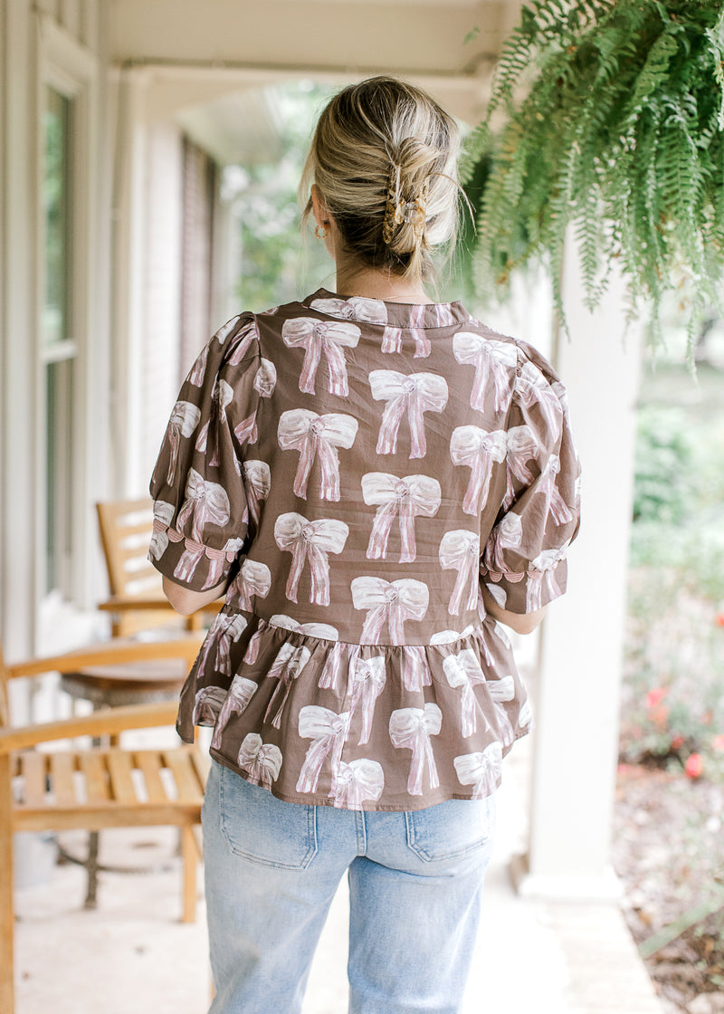 Back view of Model wearing a brown top with cream and mauve bows and short sleeves.