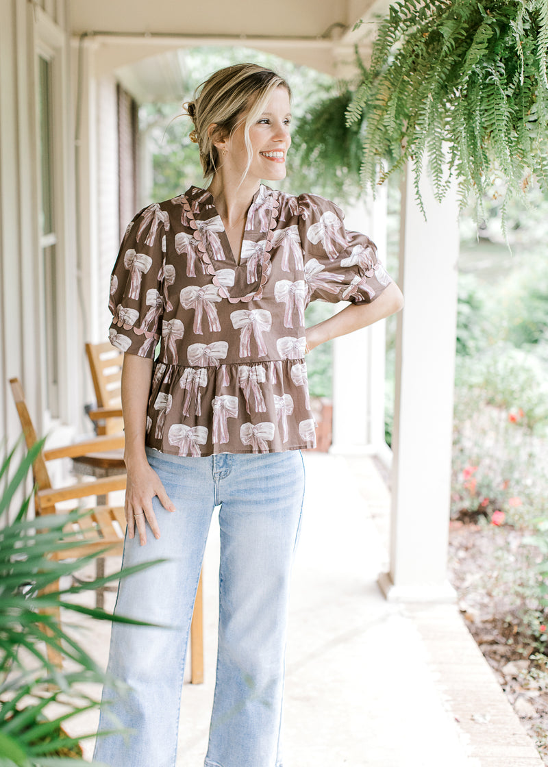 Model wearing jeans with a brown babydoll top with cream and mauve bows and short sleeves.