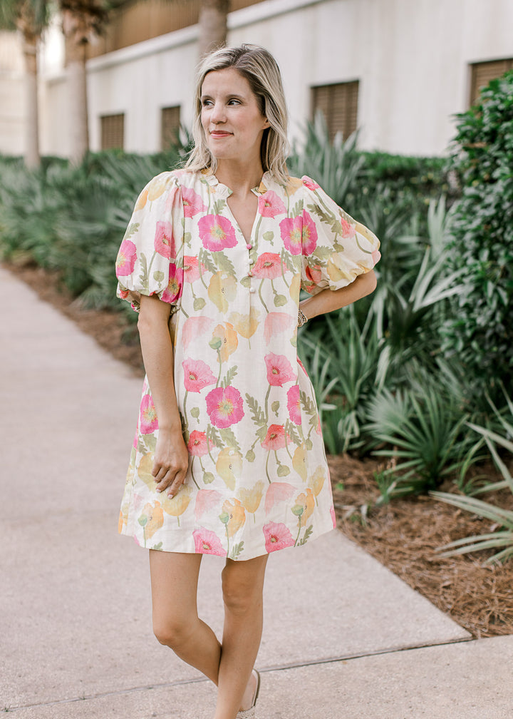 Model wearing a cream dress with pink and yellow floral, v-neck with buttons and short sleeves.