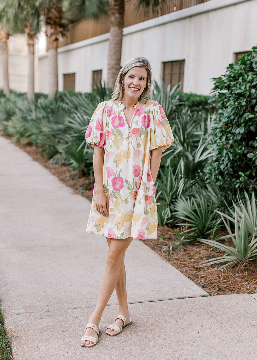 Model wearing sandals and a cream dress with floral, v-neck with buttons and bubble short sleeves.