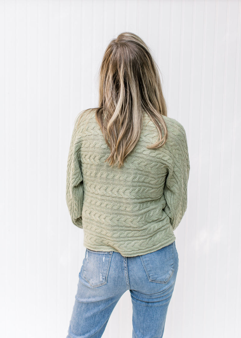 Back view of Model wearing a soft sage cable knit sweater with a mock turtle neck and long sleeves. 