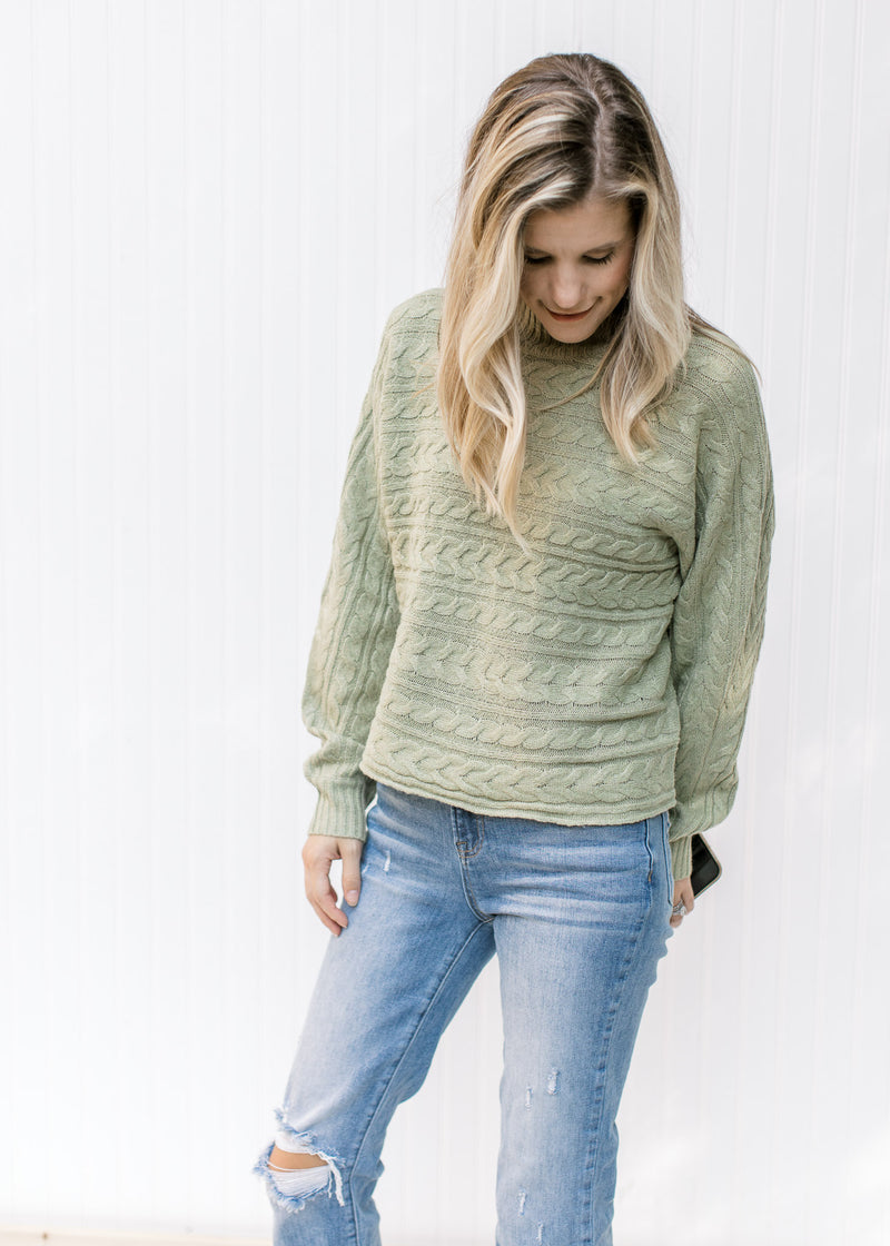 Model wearing jeans with a soft sage cable knit sweater with a mock turtle neck and long sleeves. 