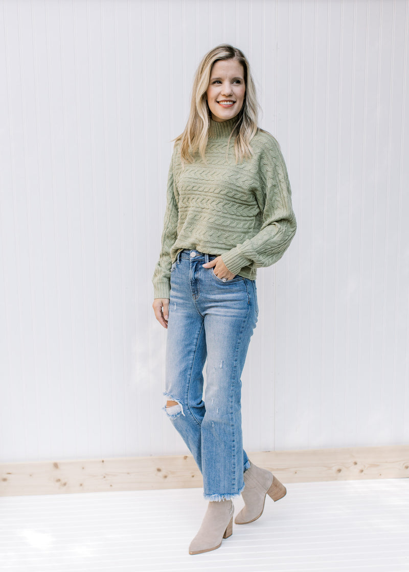 Model wearing jeans, booties and a soft sage cable knit sweater with a mock turtle neck. 