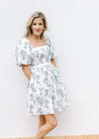 Model wearing a cream above the knee dress with pale blue floral square neck and short sleeves. 