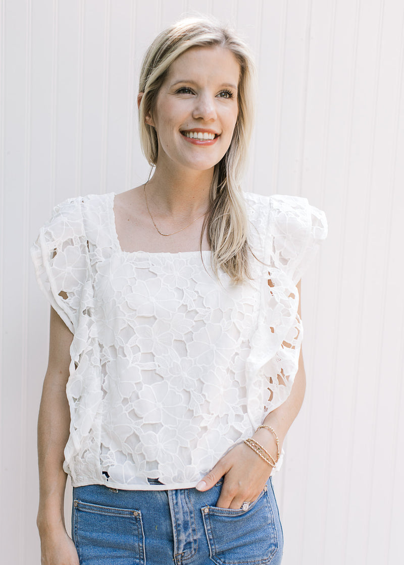 Model wearing a white top with a lace flower cut out, layered cap sleeves and a square neck. 