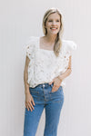 Model wearing jeans and a white top with a lace flowers, layered cap sleeves and a square neck.