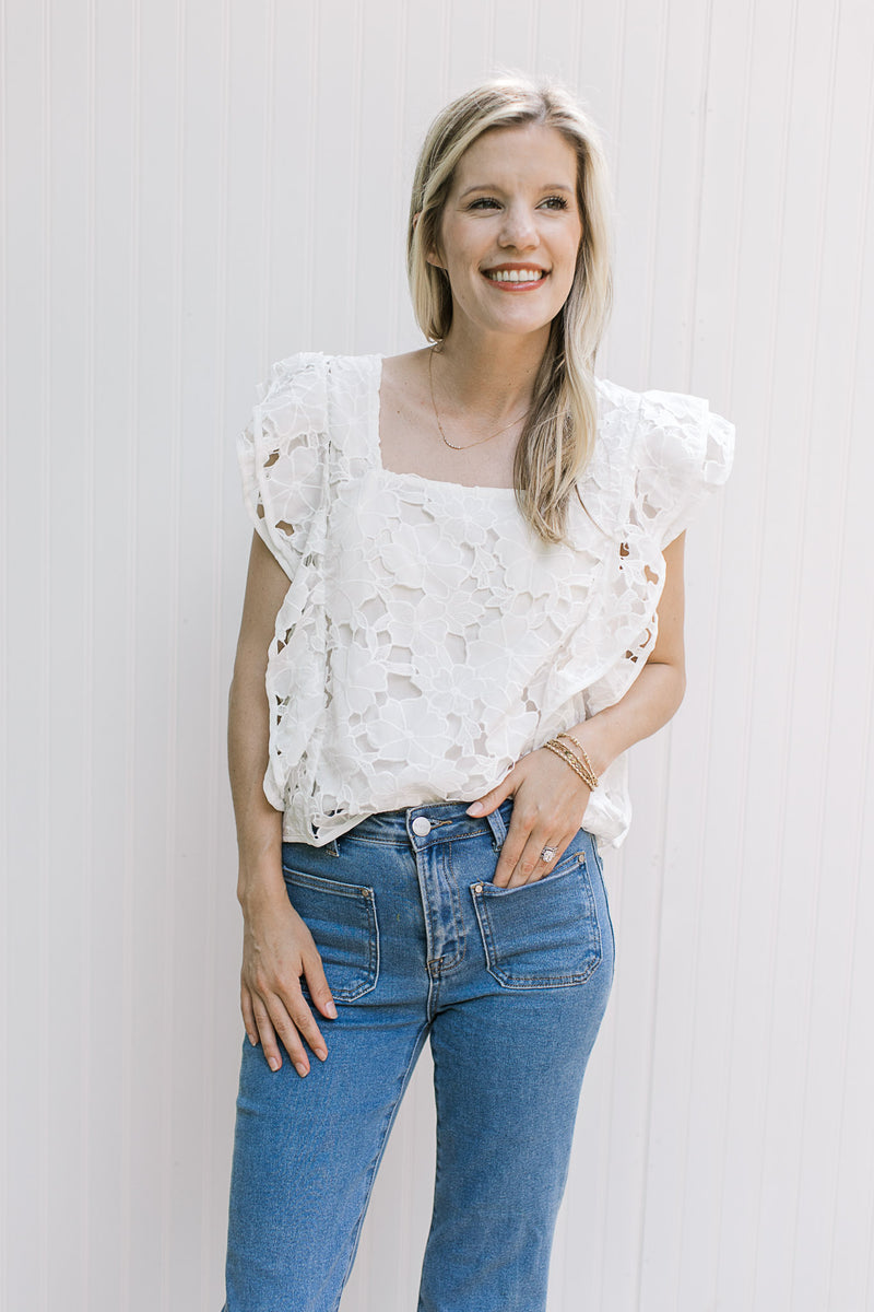 Model wearing jeans and a white top with a lace flowers, layered cap sleeves and a square neck.