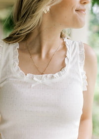 Close up of bow and ruffles at neck and sleeves of a white sleeveless ribbed top. 