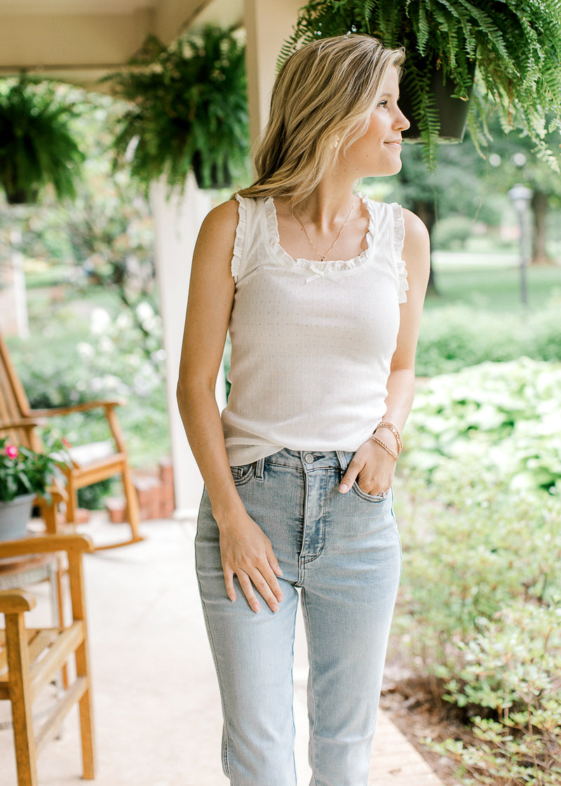 Model wearing jeans and a white ribbed top with ruffle at sleeves and neck and a bow at neck. 