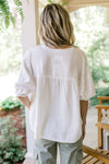 Back view of Model wearing a cool white, button up, v-neck top with short sleeves and a ruffle.