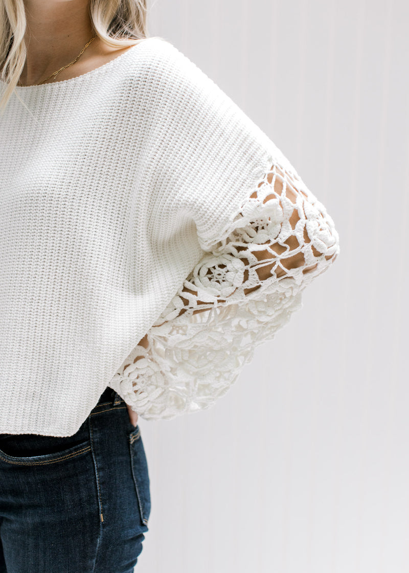 Close up of floral crochet on the sleeves of a white sweater with a crop fit and a wide neckline. 