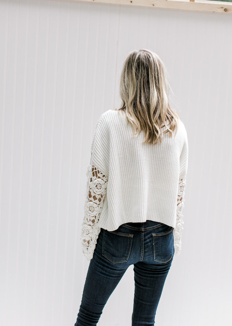 Back view of Model wearing a white top with long floral crochet sleeves, a crop fit and round neck. 