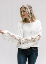 View of floral crochet detail on the sleeve of a white sweater with a crop cut and a wide neck. 