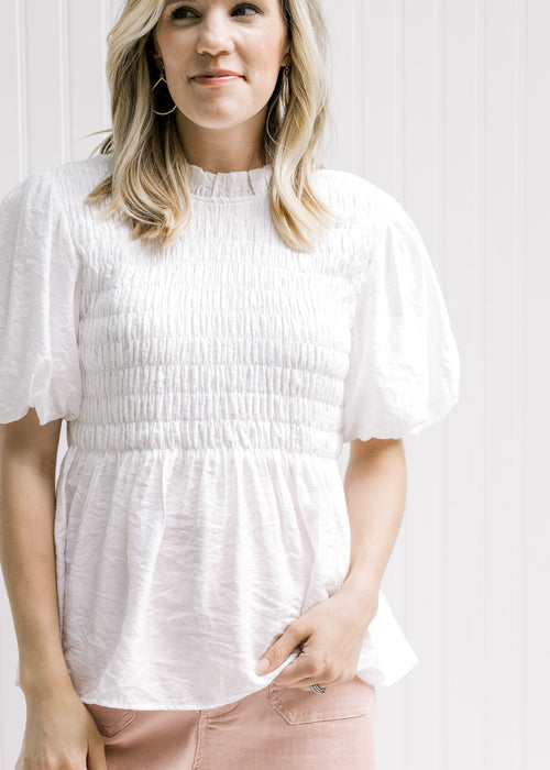 Model wearing a white top with a smocked bodice, puff sleeves and a round neck with a ruffle. 
