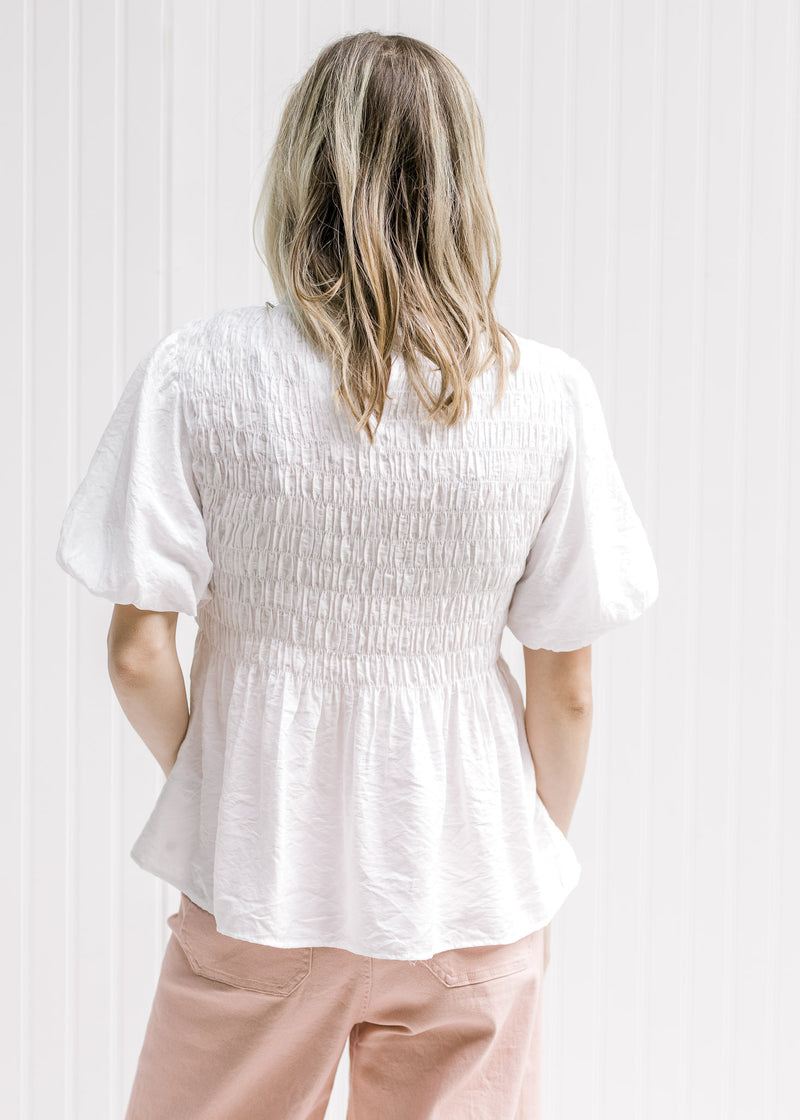 Back view of Model wearing a white top with a smocked bodice and a round neck with a ruffle. 