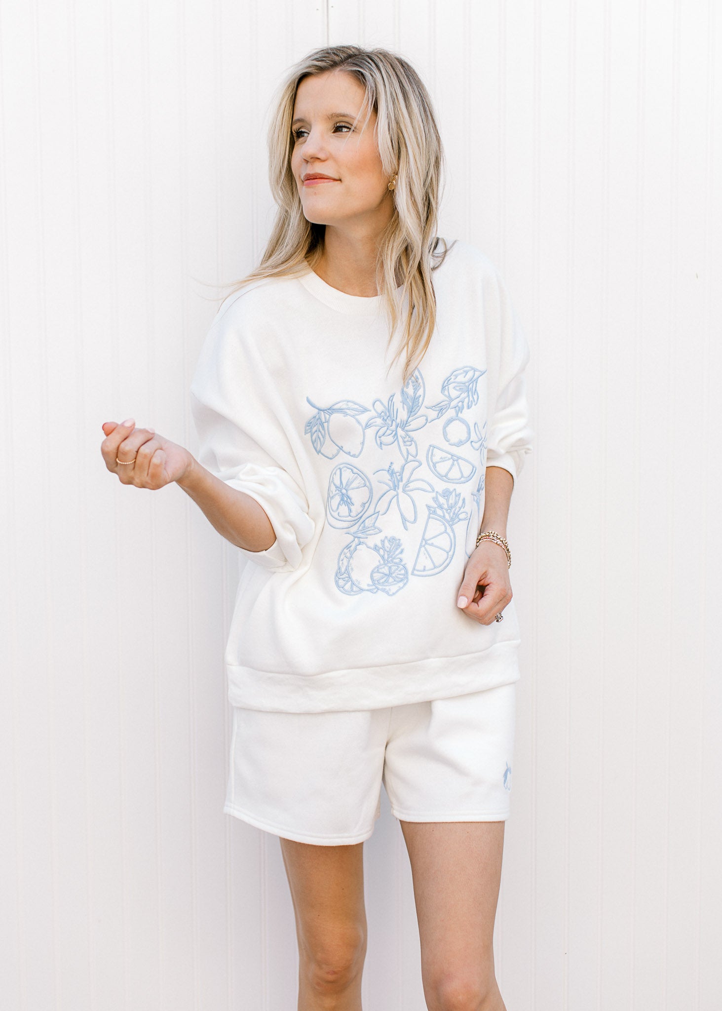 Model wearing a lightweight white sweatshirt/short set with fruit embroidered in blue. 