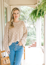 Model wearing a taupe knit sweater with embroidered daises scalloped hem and long sleeves. 