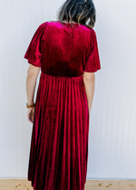 Back view of Model wearing a velvet wine midi with a pleated skirt, short sleeves and faux wrap top.