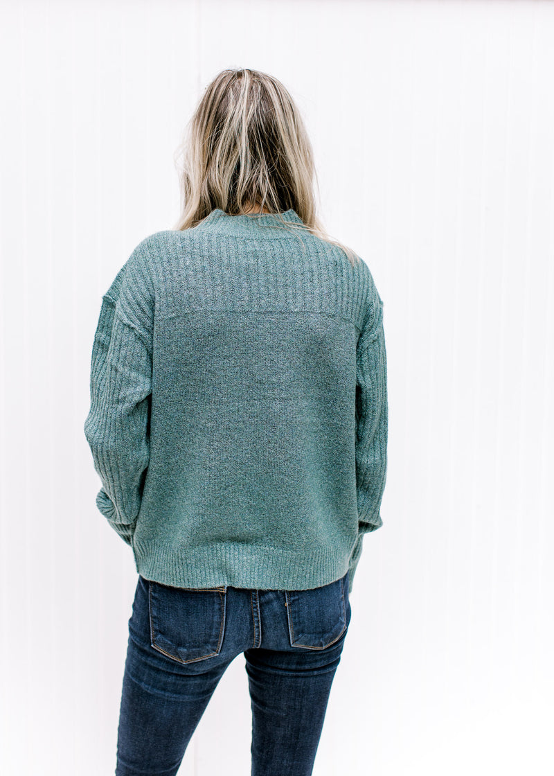 Back view of Model wearing a soft forest sweater with a mock neck, exposed hem and long sleeves.