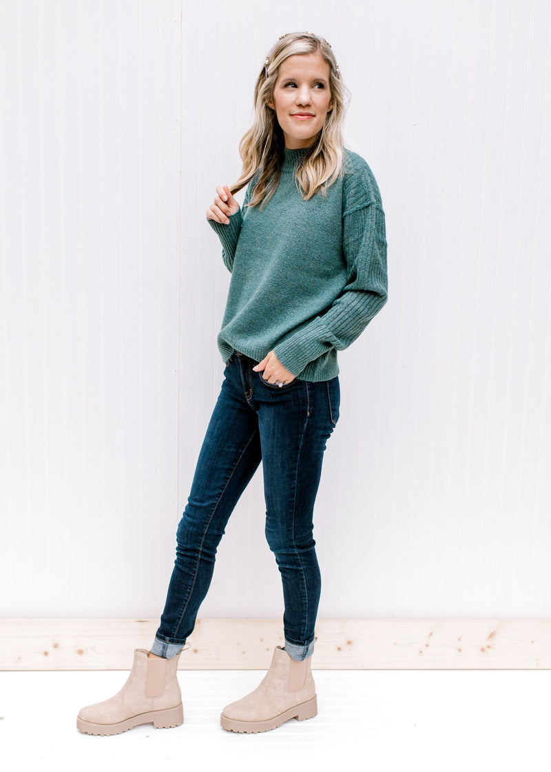Model wearing jeans, booties and a soft forest sweater with a mock neck and exposed hem. 