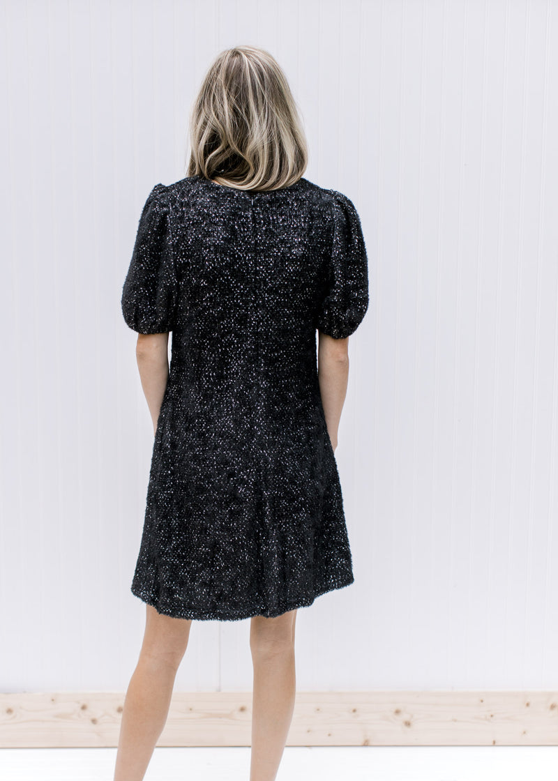 Back view of Model wearing a black dress with white speckles, bubble short sleeve and round neck.