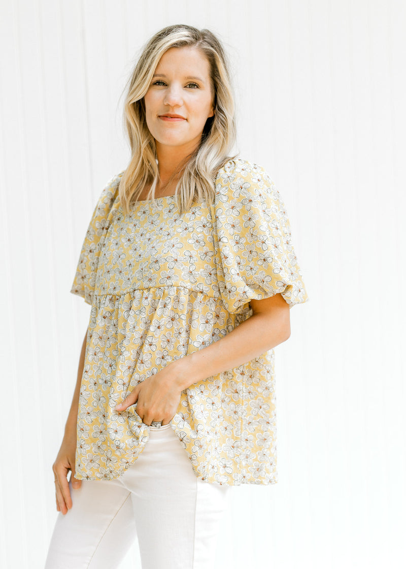 Model wearing white jeans with a mustard top with a cream floral pattern and a square neckline. 