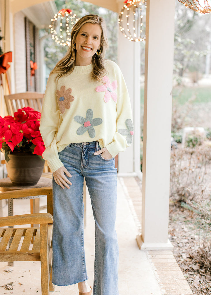 Model wearing jeans and a yellow sweater with stitched flowers, round neck and long sleeves. 