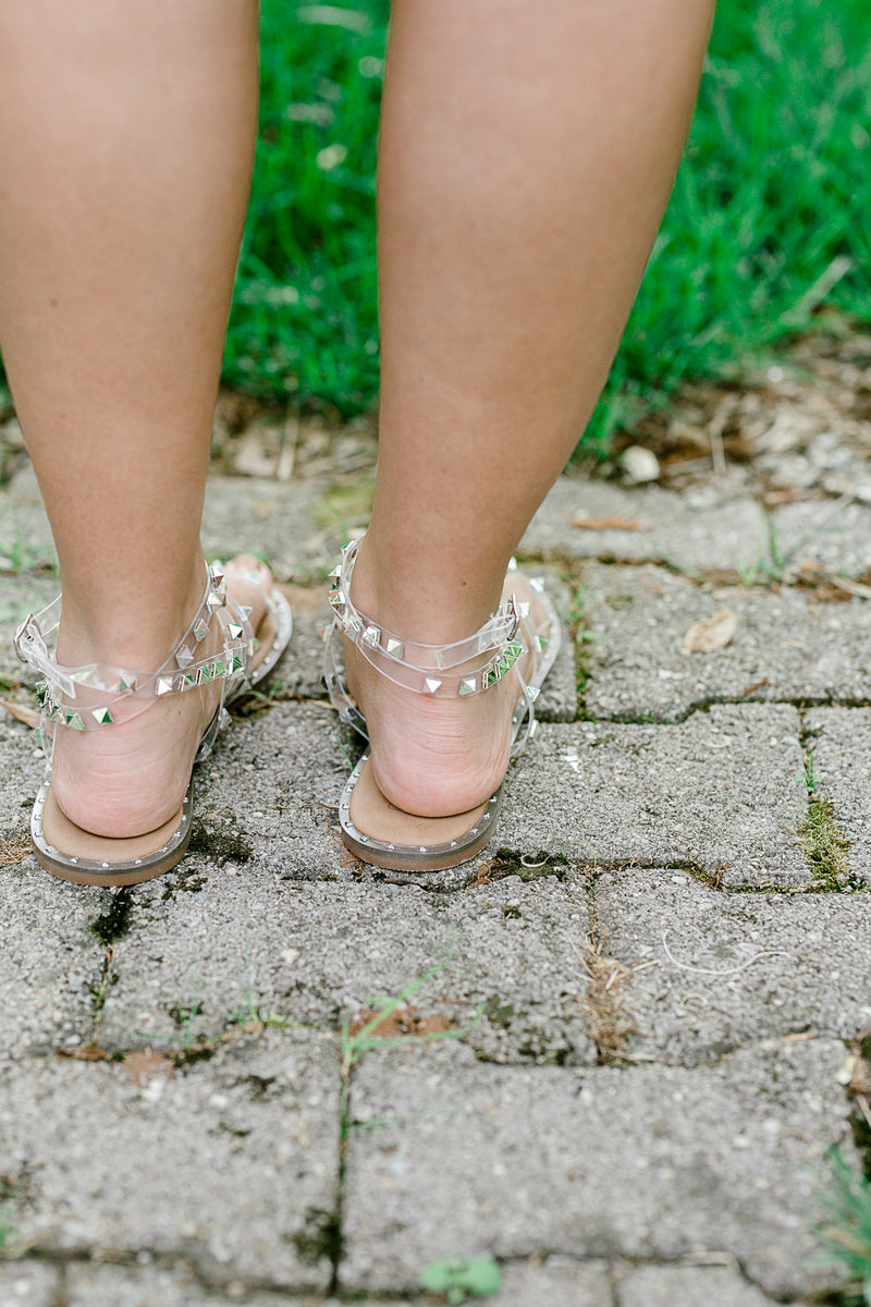 Back view of A pair of clear lucite sandals with silver stud on model.