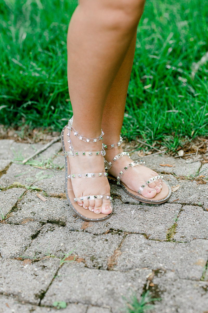 A pair of clear lucite sandals with silver stud on model on pavement.